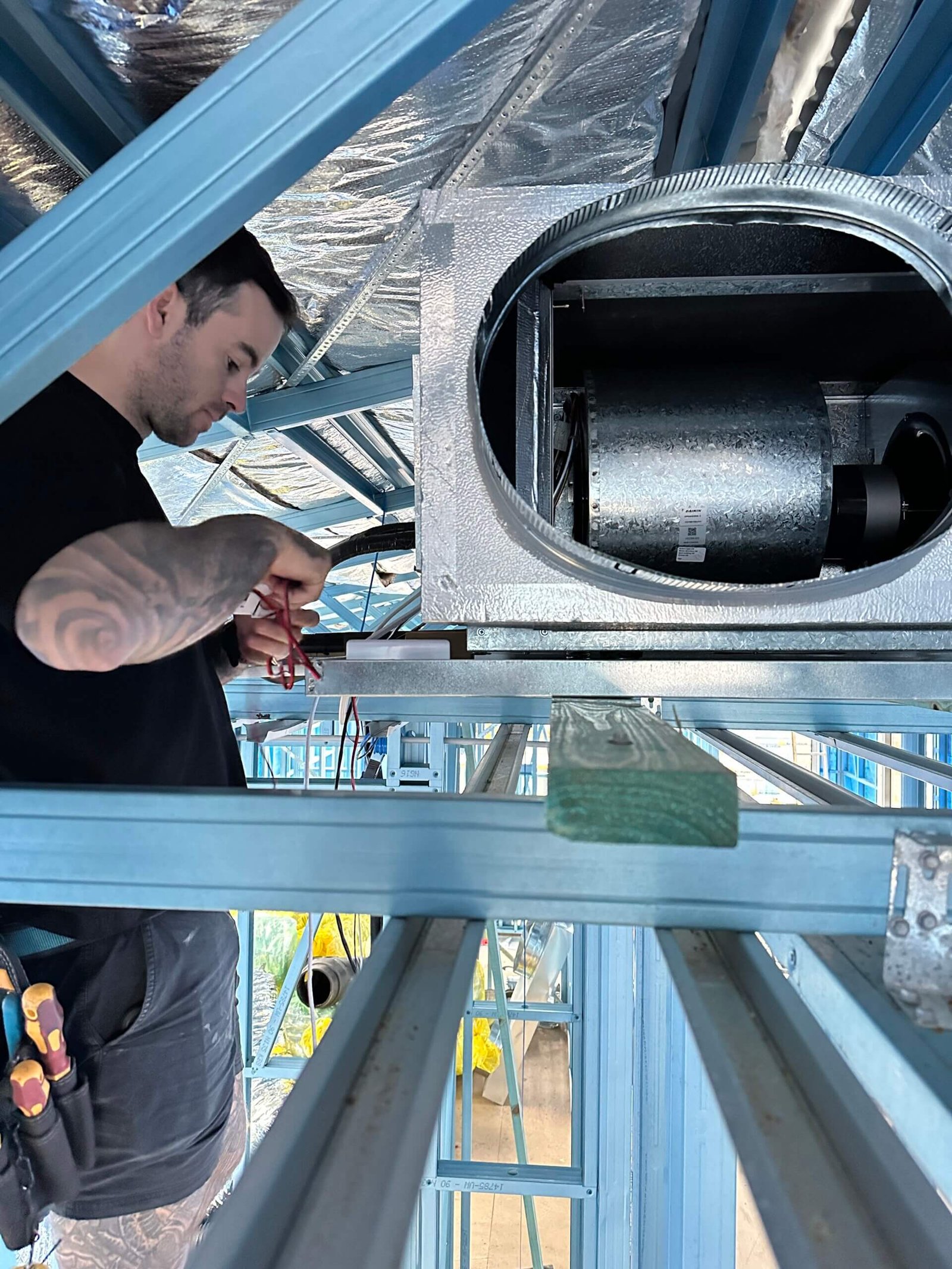 Technician performing electrical rough-in wiring on a ducted air conditioning indoor fan coil unit inside a steel frame house